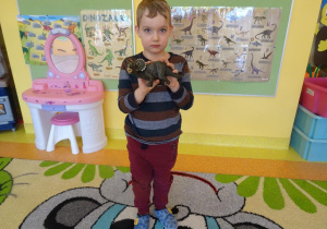 Chłopiec stoi na dywanie, w ręku trzyma figurkę dinozaura.